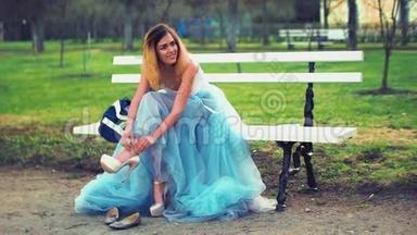 穿着银色和蓝色连衣裙的迷人女孩坐在帕克威的长凳上，穿上高跟鞋，摆出姿势，站起来。